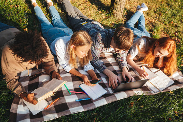 Top view of group of happy multhiethnic students doing homework together at the park, using laptop computer, laying on a grass