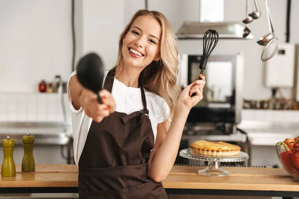 Smiling young woman chef cook in apron standing at the kitchen, holding ladle and whisk