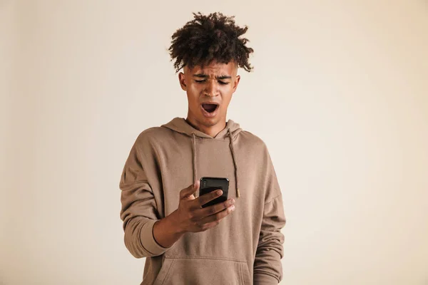 Portrait of a shocked young afro american man dressed in hoodie isolated, holding mobile phone