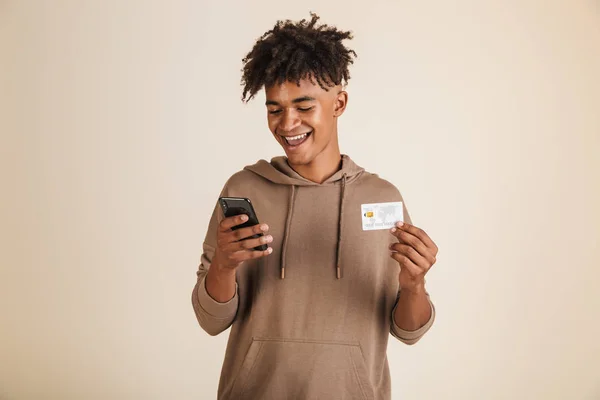 一个积极的年轻美国黑人男子的肖像身着帽衫隔离 使用手机 显示塑料信用卡 — 图库照片