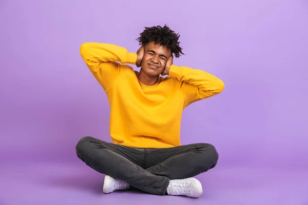 不愉快的非洲裔美国男孩的肖像坐在地板上 双腿交叉和覆盖在紫色背景的双手隔离耳朵 — 图库照片