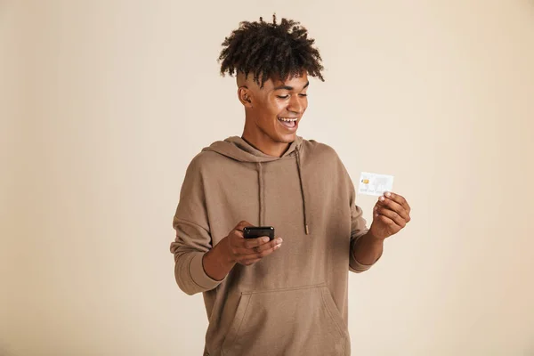 一个微笑的年轻美国黑人男子的肖像身着帽衫隔离 使用手机 显示塑料信用卡 — 图库照片#