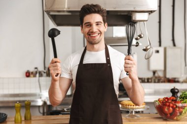 Mutlu genç adam şef yemek önlük daimi mutfak at holding kepçe ve çırpma teli