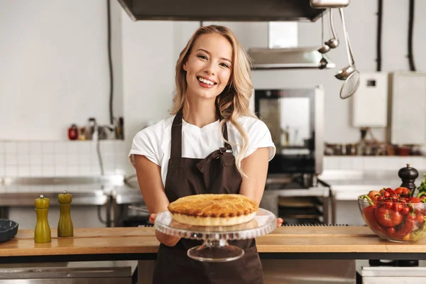 Smiling young woman chef cook in apron standing at the kitchen, showing tasty pie