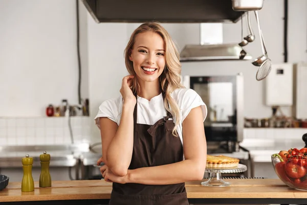 Smiling young woman chef cook in apron standing at the kitchen, looking at camera