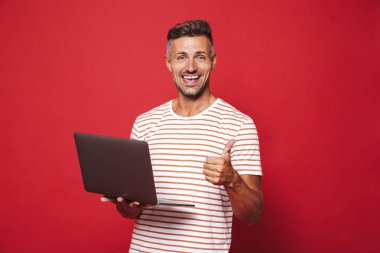 Gülümseyen ve gri laptop izole kırmızı arka plan üzerinde tutarak çizgili tişört iyimser tıraşsız adam