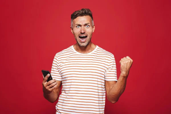 Portrait of an excited man standing over red background, holding mobile phone