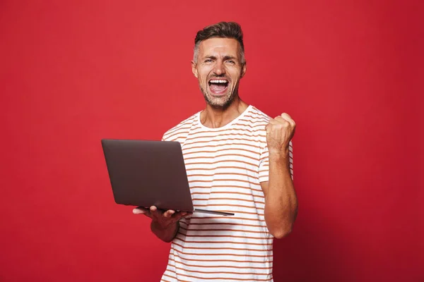 Image of successful guy in striped t-shirt smiling and holding gray laptop isolated over red background