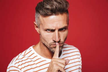 Çizgili tişört holding işaret parmağı ağzında izole kırmızı arka plan üzerinde sıkı bakışla kendine güvenen adam