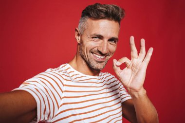 Gülümseyen ve selfie, kırmızı arka plan üzerinde izole çekerken ok işaret gösteren çizgili tişört genç adam
