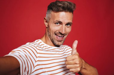 Gülümseyen ve başparmak süre alarak selfie izole kırmızı arka plan üzerinde gösterilen çizgili tişört neşeli adam