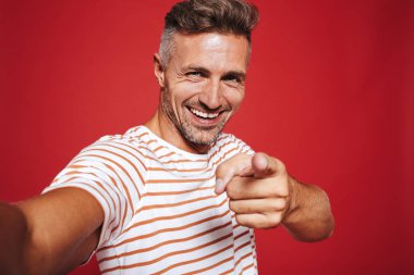 Gülümseyen ve sana selfie kırmızı arka plan üzerinde izole çekerken parmakla çizgili tişört neşeli adam
