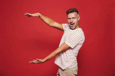 Excited man in striped t-shirt gesturing and demonstrating size with copyspace isolated over red background clipart