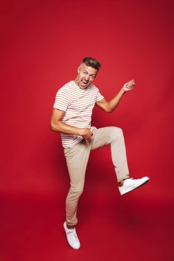 Genç adam fotoğrafı tam uzunlukta çığlık ve görünmez gitar üzerinde kırmızı arka plan izole çalmaya t-shirt çizgili