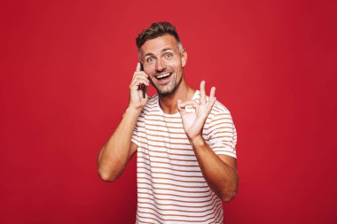 Kırmızı arka plan üzerinde duran, cep telefonu üzerinde konuşurken, ok gösterilen heyecanlı bir adam portresi