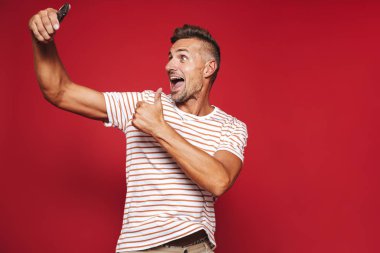 Kırmızı arka plan üzerinde duran, bir selfie alarak, başparmak gösterilmesini neşeli bir adam portresi