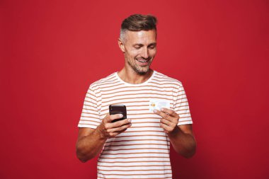 Kırmızı arka plan üzerinde duran, cep telefonu tutan, kredi kartı gösteren gülümseyen bir adam portresi