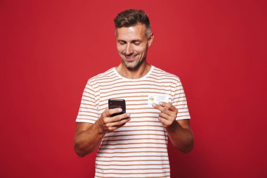 Kırmızı arka plan üzerinde duran, cep telefonu tutan, kredi kartı gösteren mutlu bir adam portresi