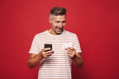 Kırmızı arka plan üzerinde duran, cep telefonu tutan, kredi kartı gösteren heyecanlı bir adam portresi
