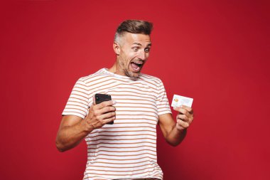 Kırmızı arka plan üzerinde duran, cep telefonu tutan, kredi kartı gösteren neşeli bir adam portresi