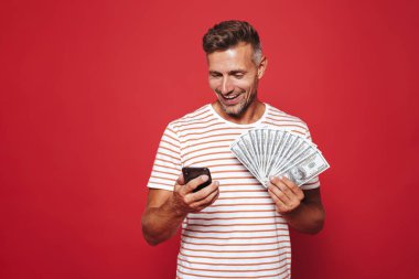 Kırmızı arka plan üzerinde duran, cep telefonuyla, para banknot gösterilen mutlu bir adam portresi