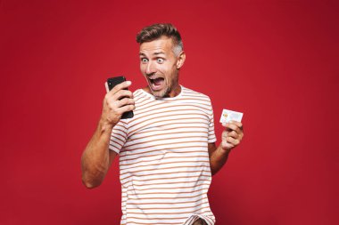Kırmızı arka plan üzerinde duran, cep telefonu tutan, kredi kartı gösteren şok bir adam portresi