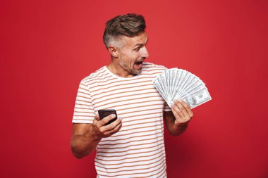 Kırmızı arka plan üzerinde duran, cep telefonuyla, para banknot gösterilen mutlu bir adam portresi
