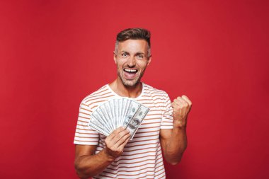 Görüntü gülümseyen ve izole kırmızı arka plan üzerinde nakit para hayranı tutan çizgili tişört memnun insanın
