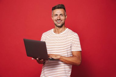 Görüntü bir süre tutarak ve izole kırmızı arka plan üzerinde dizüstü kullanarak şeritli t-shirt gülümseyen içinde beyaz adam 30'lu