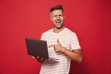 Görüntü gülümseyen ve gri laptop izole kırmızı arka plan üzerinde tutarak çizgili tişört çekici adamın