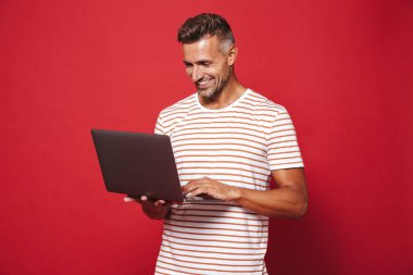 Görüntü gülümseyen ve gri laptop izole kırmızı arka plan üzerinde tutarak çizgili tişört esmer adamın