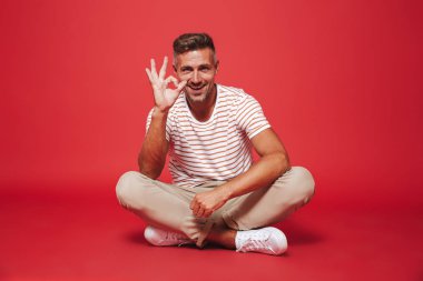 Bacaklar ile katta otururken çizgili gömlek gülümseyen neşeli adam 30'lu fotoğraf izole kırmızı arka plan üzerinde çarpı işareti