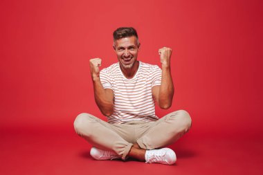 Yerde bağdaş ile izole kırmızı arka plan üzerinde otururken çizgili tişört kenetlenmesi içinde yakışıklı adam 30'lu görüntüsünü yumruklar