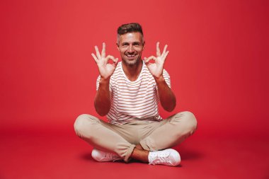 Bacaklar ile katta otururken çizgili gömlek gülümseyen içinde Avrupa adam 30'lu görüntü izole kırmızı arka plan üzerinde çarpı işareti