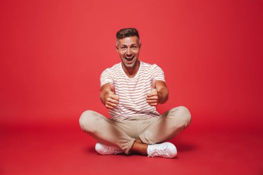 Bacaklar ile katta otururken çizgili gömlek gülümseyen içinde çekici bir adam 30'lu görüntü izole kırmızı arka plan üzerinde çarpı işareti