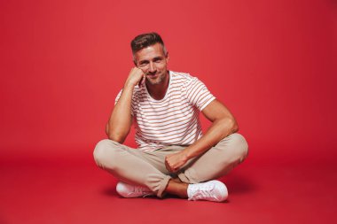 Bacaklar ile katta otururken çizgili gömlek gülümseyen yakışıklı adam 30'lu görüntü izole kırmızı arka plan üzerinde çarpı işareti