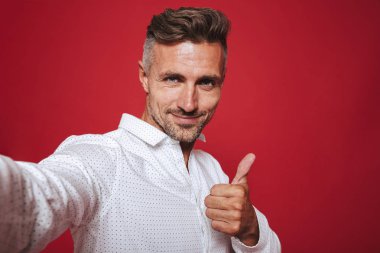 Yakışıklı esmer 30'lu yaşlarda olan adam anız beyaz gömlekli selfie fotoğraf ve gösteren başparmak kırmızı arka plan üzerinde izole alarak