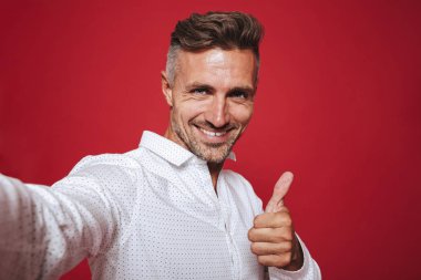 İyimser esmer 30'lu yaşlarda olan adam anız beyaz gömlekli selfie fotoğraf ve gösteren başparmak kırmızı arka plan üzerinde izole alarak