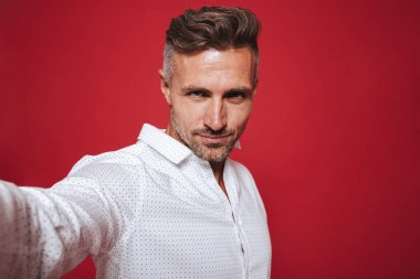 Yakışıklı sakallı adam 30'lu yaşlarda beyaz gömlek gülümseyen ve selfie fotoğrafta kırmızı arka plan üzerinde izole