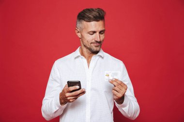 Kredi kartı ve kırmızı arka plan üzerinde izole smartphone holding Beyaz tişörtlü adam gülümsüyor, fotoğraf