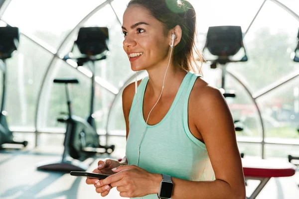 Happy sports woman in earphones holding smartphone and looking away while being in gym