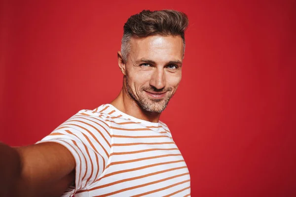 Beautiful guy 30s in striped t-shirt smiling while taking selfie photo isolated over red background