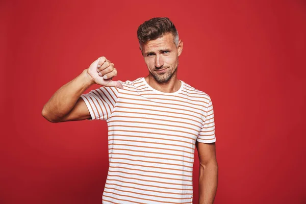 Confused man in striped t-shirt showing thumb down and expressing dislike isolated over red background