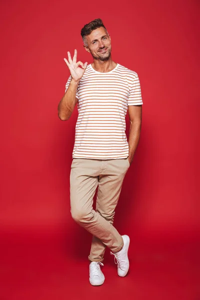 Full length photo of brunette man in striped t-shirt smiling and showing ok sign isolated over red background
