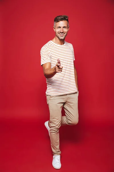 Full length photo of positive man in striped t-shirt smiling and showing thumb up isolated over red background