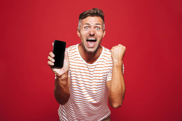Portrait of an excited man standing over red background, showing mobile phone, celebrating