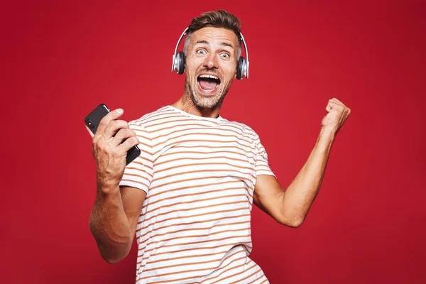 Portrait of an excited man standing over red background, listening to music with headphones, holding mobile phone