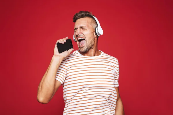 一个开朗的男人的肖像站在红色背景 听音乐与耳机 手持手机 — 图库照片