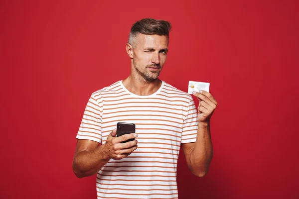 一个男人的肖像站在红色背景 手持手机 显示信用卡 — 图库照片
