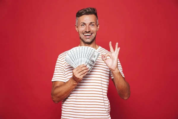 Image of rich guy in striped t-shirt smiling and holding fan of money banknotes isolated over red background